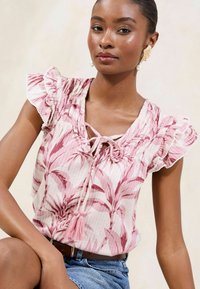 Floral patterned pink blouse with ruffle sleeves, a tie neckline, and tassel detail. Lightweight fabric features subtle texture.