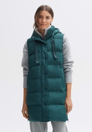 Gepolsterte türkisfarbene Weste mit Kapuze, die über einen Knopfverschluss, verstellbare Träger und eine gesteppten Textur verfügt. Getragen über einem grauen Pullover.