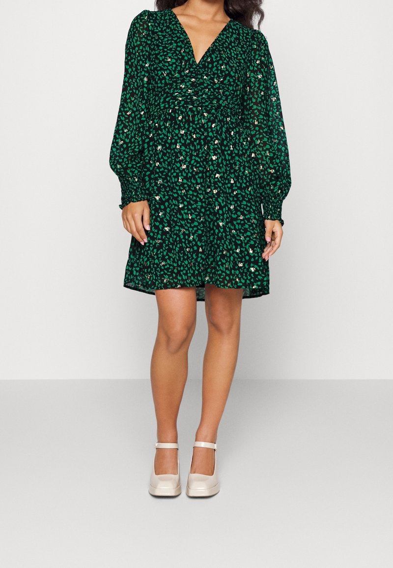 Kvinna i en grön och svart blommig klänning med V-ringning och långa pufärmar, tillsammans med beige Mary Jane-skor, stående mot en neutral bakgrund.