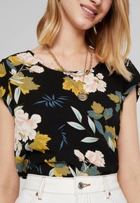 Blusa nera floreale con maniche corte, caratterizzata da fiori gialli, rosa e blu. Tessuto morbido con collo rotondo e delicato motivo floreale.