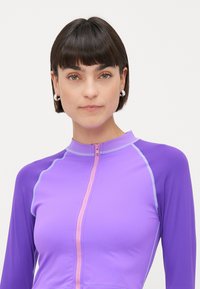 Sieviete ar īsiem tumšiem matiem, uzvilkusi sudraba gredzenveida auskarus un violetu sporta zip-up kreklu ar kontrastējošām šuvēm uz neitrāla fona.