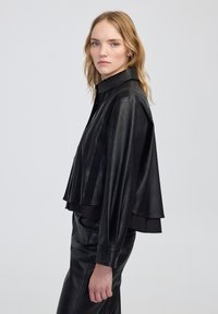 Czarna skórzana kurtka o kroju cropped z kołnierzem na guziki i opadającymi ramionami. Posiada gładką fakturę i warstwowy design.