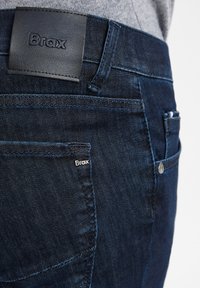 Donkerblauwe denim jeans met een gladde textuur, met een leren taillebandlabel dat is ingebrand met "Brax" en een klein zijlogo-tag.