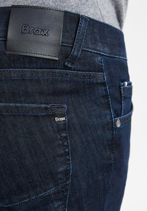 Dunkelblaue Jeans aus Denim mit glatter Textur, ausgestattet mit einem Lederbundetikett, das mit "Brax" geprägt ist, sowie einem kleinen seitlichen Logoetikett.
