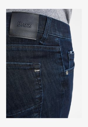 Dunkelblaue Jeans aus Denim mit glatter Textur, ausgestattet mit einem Lederbundetikett, das mit "Brax" geprägt ist, sowie einem kleinen seitlichen Logoetikett.