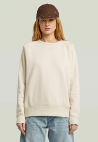 Light beige sweatshirt with a crew neck and raglan sleeves, paired with a brown cap featuring a small logo. Soft, casual fabric.