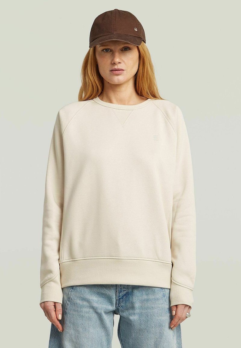 Light beige sweatshirt with a crew neck and raglan sleeves, paired with a brown cap featuring a small logo. Soft, casual fabric.