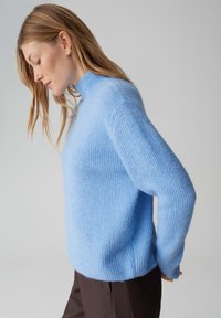 Pull en laine bleu clair avec un col montant, une texture côtelée et une coupe légèrement oversize. Le modèle porte un pantalon marron foncé, debout de profil.