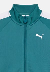 Τζελ αθλητικό μπουφάν με φερμουάρ σε τιρκουάζ χρώμα, με λευκές ρίγες στους ώμους και λευκό λογότυπο Puma στο στήθος, εμφανιζόμενο από τον γιακά μέχρι τη μέση του κορμού.