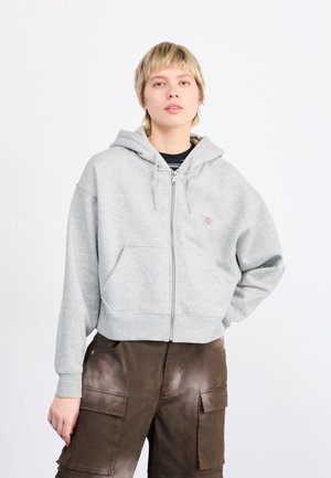 Sudadera con cremallera gris, con un bolsillo frontal, capucha y puños acanalados; presenta un pequeño logo en el pecho. Lleva puesta con pantalones cortos de cargo marrones.