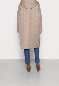 Cappotto imbottito beige con cappuccio, caratterizzato da un design lungo e dettagli con spacco laterale. Indossato sopra jeans blu e stivali con tacco marroni.