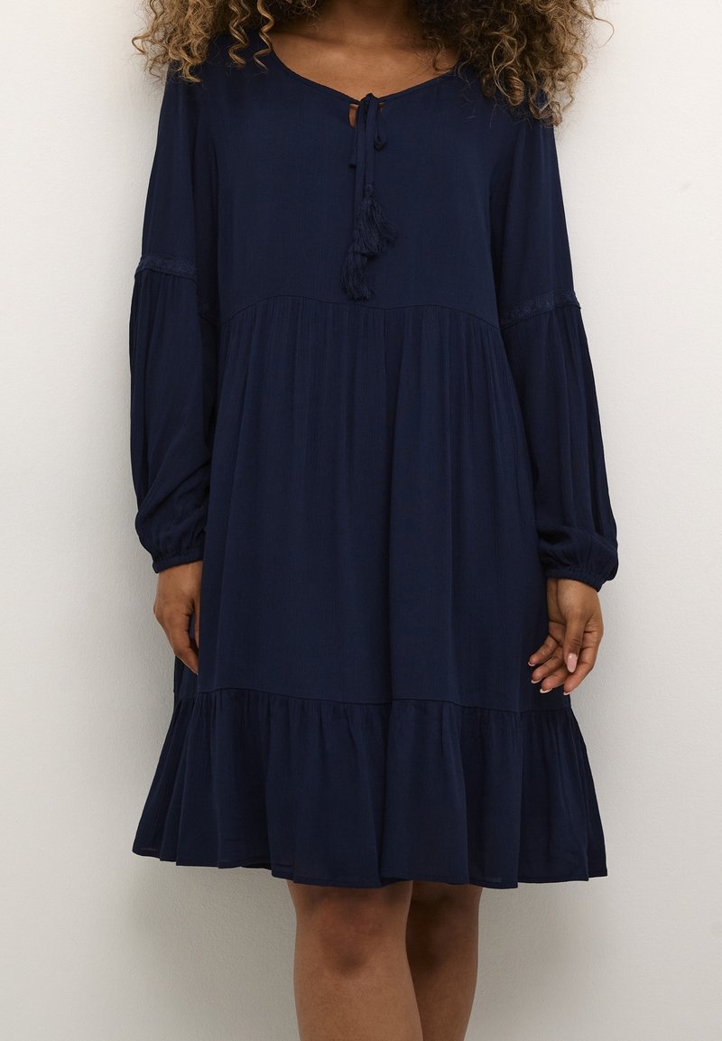 Marinblå klänning med rund halsringning, knytdetalj, långa puffärmar och en flerlagers volangkant. Tillverkad av lätt tyg med en mjuk textur.