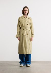 Femme debout face à l'avant, portant un trench-coat beige ceinturé, un jean bleu à jambes larges, et des baskets argentées sur un sol gris.