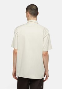 Camicia a maniche corte in tessuto beige chiaro, con colletto standard e taglio dritto, caratterizzata da una texture liscia e minimi dettagli.