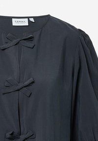 Dunkelgraues Blusenshirt mit Rundhalsausschnitt, puffärmeligen Ärmeln und dekorativen Stoffschleifen entlang der vorderen Verschlussleiste. Weiche Textur mit einem dezenten Glanz.