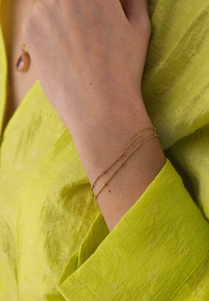 Wrist wearing two delicate gold chain bracelets with small beads over a yellow textured long-sleeve garment.