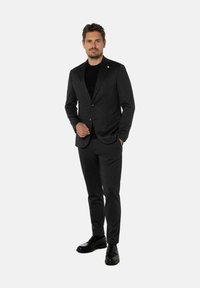 Man in een donkergrijs pak en zwarte schoenen staat met een hand in zijn zak tegen een effen lichte achtergrond.