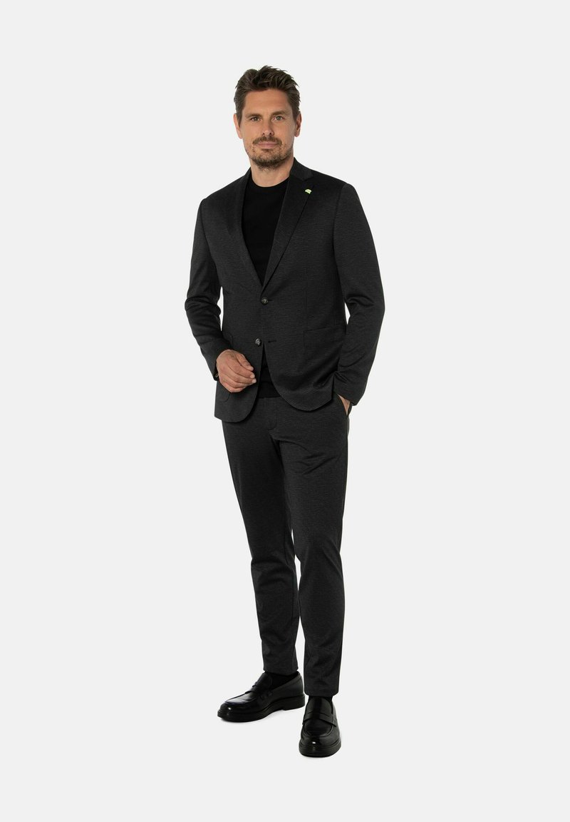 Man in een donkergrijs pak en zwarte schoenen staat met een hand in zijn zak tegen een effen lichte achtergrond.