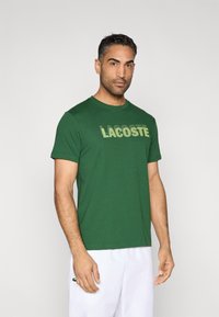Camiseta verde de manga corta con la palabra "LACOSTE" impresa en un tono más claro. Tela suave, cuello redondo, corte relajado, detalles mínimos.