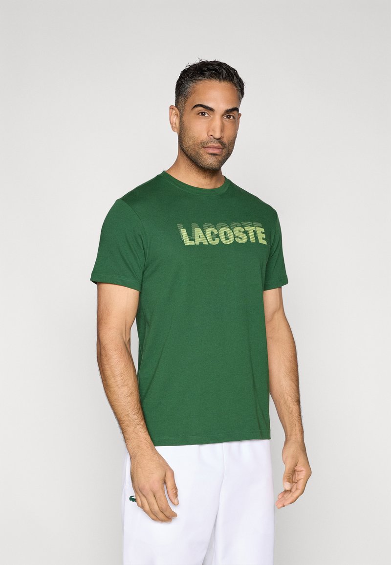 Camiseta verde de manga corta con la palabra "LACOSTE" impresa en un tono más claro. Tela suave, cuello redondo, corte relajado, detalles mínimos.