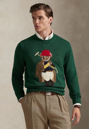 Zelený pletený sveter s motívom medveďa v červenej čiapke s golfovou palicou. Textúrované pletenie a rebrované manžety, v kombinácii s kockovanými nohavicami.
