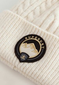 Jasnobeżowy dzianinowy beret z grubym prążkowanym wzorem oraz okrągłą naszywką z haftem przedstawiającym góry i napisem "SUPERDRY".