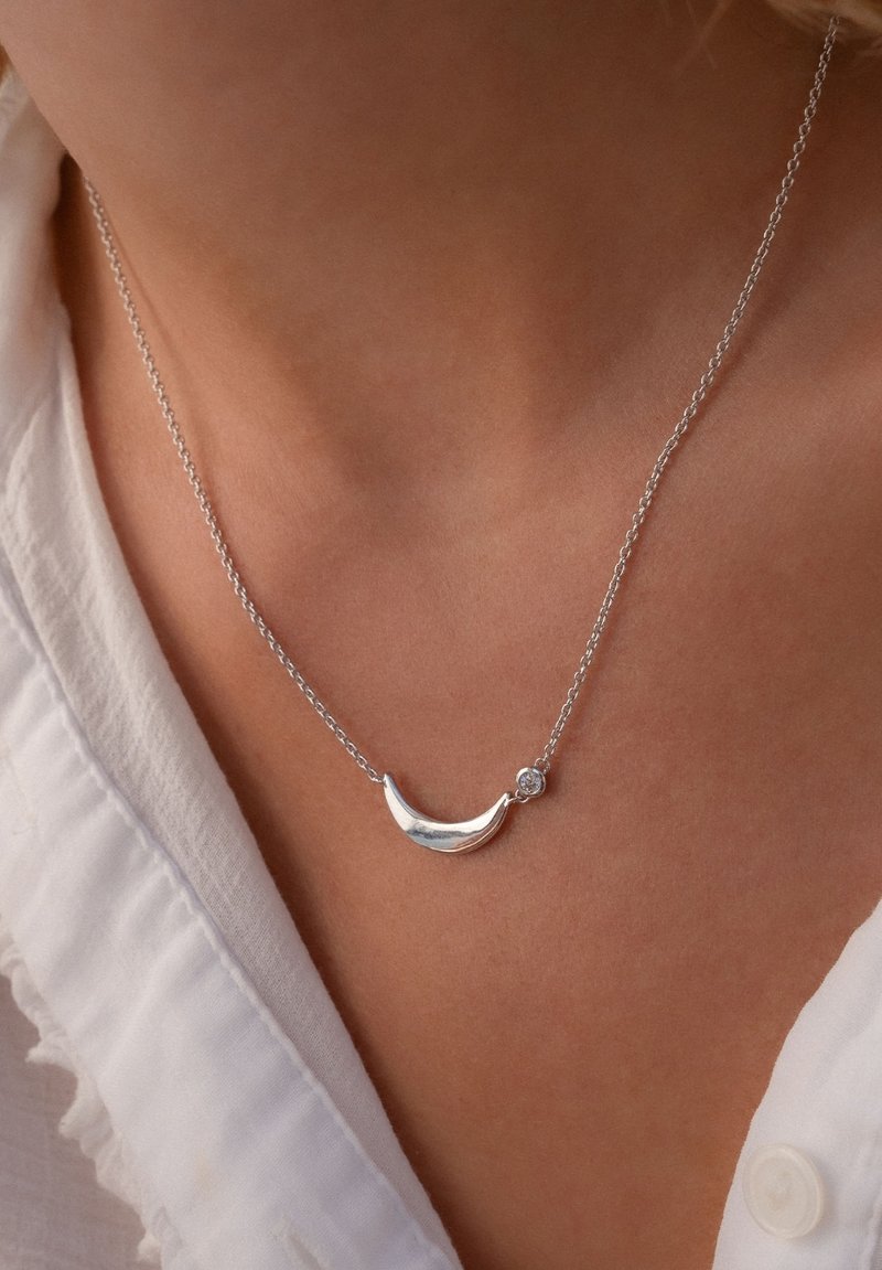 Silberne Halskette mit einem Halbmond-Anhänger mit polierter Oberfläche, begleitet von einem kleinen klaren Edelstein, an einer zarten Kette.