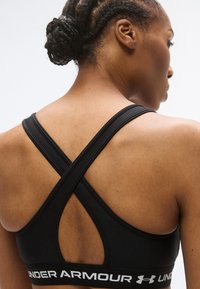 Μαύρο αθλητικό σουτιέν με σχέδιο διασταυρούμενης πλάτης, με απαλό υλικό και ελαστική ταινία με το κείμενο "UNDER ARMOUR" σε λευκό χρώμα.