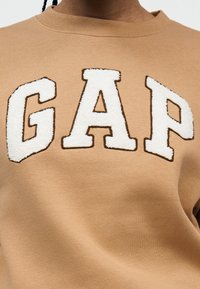 Gros plan d'un sweat-shirt beige avec de grandes lettres texturées blanches formant le mot "GAP" sur la poitrine.