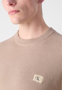 Maglione beige in maglia con motivo testurizzato, collo rotondo a coste e una piccola etichetta con il logo "CK" nell'angolo in basso a sinistra.