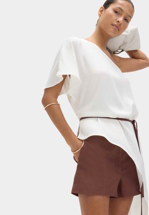 Kvinde iført en hvid asymmetrisk kortærmet bluse og brune shorts med høj talje, accessoriseret med guldbøjler og holder en hvid clutch.