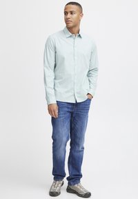 Camisa de botones azul claro con cuello, combinada con vaqueros azul y zapatillas deportivas grises. La tela parece suave y casual.