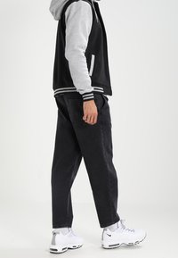 Giacca bomber nera e grigia con polsini a righe, abbinata a pantaloni neri larghi e sneakers bianche con accenti grigi.