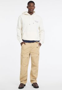 Kremowa bluza z kapturem z przednią kieszenią i graficznym nadrukiem, w połączeniu z beżowymi spodniami cargo z wieloma kieszeniami. Czekoladowe skórzane buty.