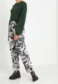 Pull en tricot vert et pantalon cargo gris camouflage, avec des poches, associés à des bottes noires à lacets et à semelles épaisses.