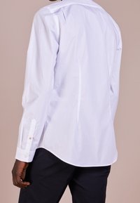 Witte blouse met lange mouwen, afgeronde zoom, knoopsluiting bij de manchetten met contrasterende knopen, en een gladde, lichte stof.