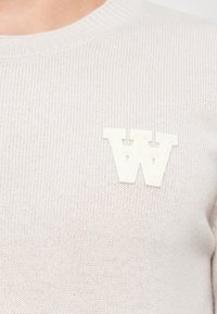 Svetlý béžový pletený sveter s rebrovanou textúrou a výraznou bielou nášivkou s logom "W" na hrudi.