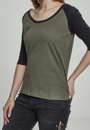 Femme portant un top vert et noir à manches trois-quarts associé à un jean noir avec un détail lacé sur la cuisse.