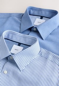 Deux chemises habillées pour hommes pliées avec col, une bleu clair uni et une blanche à rayures bleues, toutes deux étiquetées "NEXT".