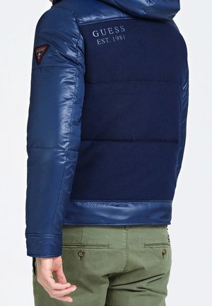 Marineblaue Daunenjacke mit glänzender, gepolsterter Oberfläche und Strickstoff auf der Rückseite. Mit einem Logo auf der Schulter und geripptem Saum.