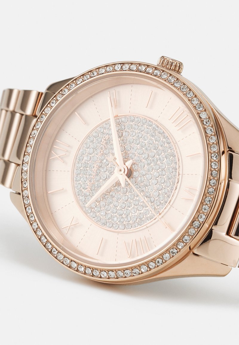 Złoty różowy zegarek z kryształowymi akcentami na bezel i tarczy. Posiada świecące kamienie oraz rzymskie cyfry i metalowe wskazówki.