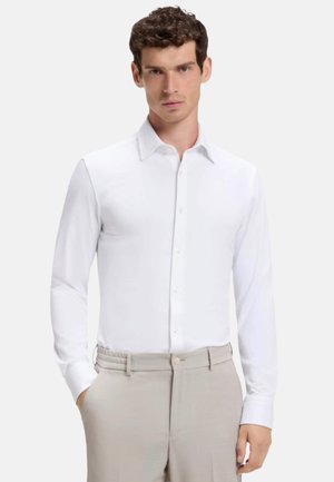 Man met kort krullend bruin haar draagt een wit shirt met lange mouwen en knopen en beige broek, staat met één hand in zijn zak.