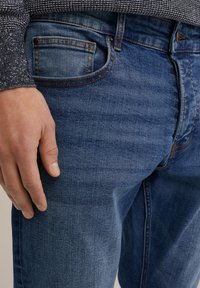 Zilā denima bikses ar izlaižamu mazgāšanu, ar oranžiem šuvju, piecām kabatām un tuvplānu ar roku, kas atpūšas uz augšstilba.