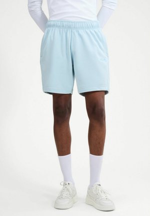 Person trägt hellblaue Sportshorts, ein weißes Langarmshirt, weiße Kniestrümpfe und weiße Turnschuhe und steht mit den Händen in den Taschen.