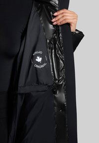 Schwarze Daunenjacke mit glänzender Innenauskleidung, mit einem Logo auf der linken Seite. Das Außenmaterial ist glatt mit einem eleganten Design.