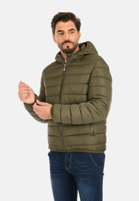 Uomo che regola la manica di una giacca puffer verde oliva con cappuccio, indossando jeans blu, in piedi contro uno sfondo chiaro e semplice.