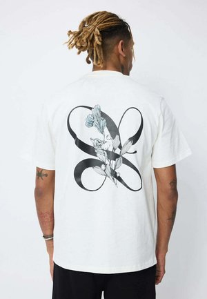 Homme avec des dreadlocks portant un T-shirt blanc orné d'une grande boucle abstraite noire et d'un motif floral bleu et blanc au dos.