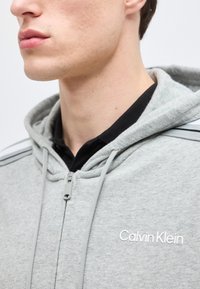 Ung person iført en lys grå glidelåshoodie med "Calvin Klein"-logo, over en svart skjorte med krage, sett fra siden.