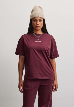 Vrouw draagt een beige gebreide muts, een bordeauxrood T-shirt met korte mouwen met kleine witte tekst, bijpassende broek, zilveren kettingen en ringen tegen een effen lichte achtergrond.