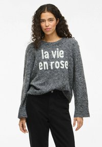 Eine Frau mit lockigem, dunklem Haar trägt einen grauen Pullover mit dem Aufdruck "la vie en rose" und schwarze, hoch taillierte Hosen, während sie vor einem weißen Hintergrund steht.